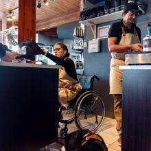 Woman in a wheelchair working behind the counter of a coffee shop and serving a customer with her service dog lying on the floor.