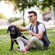 Un jeune homme aveugle est assis dans un parc municipal et caresse son chien-guide d'une main pendant qu'il tient sa canne blanche de l'autre main.