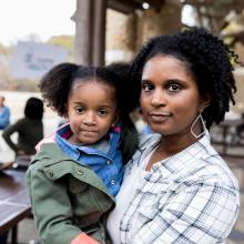 Young woman holds daughter and looks at camera with urban outdoor health clinic in background