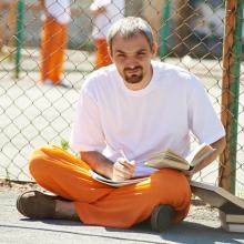 Prison inmate sitting on the ground writing in notebook and holding book