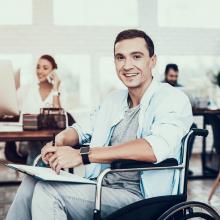 Disabled man in wheelchair at work in an office with coworkers in background. Accessibility
