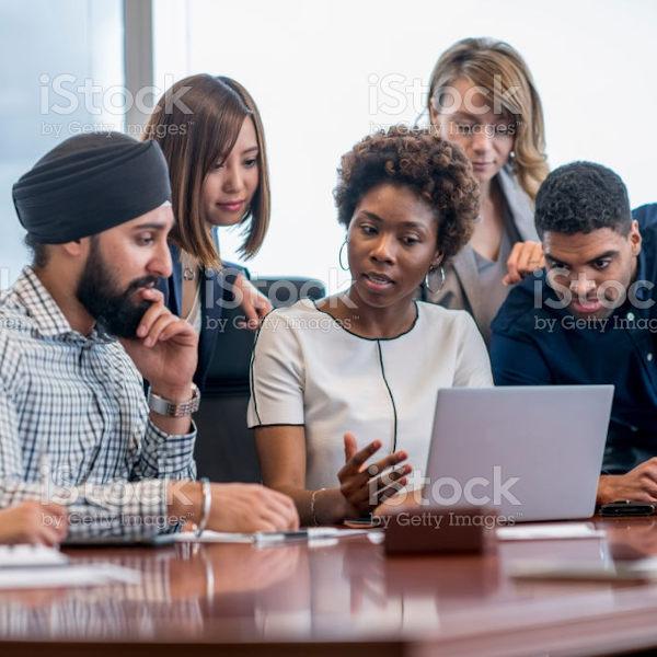 A diverse group of business people gather around a laptop in a modern office and discuss what they see.