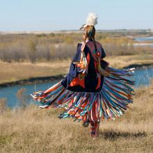 First Nations woman performing a Fancy Shawl Dance in a grass field with a river background