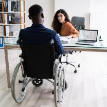 Une femme assise derrière un bureau discute avec un collègue en fauteuil roulant devant elle. Accessibilité