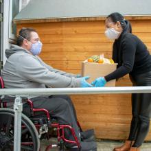 Man in a wheelchair wearing a mask and medical gloves accepts a box of groceries from a woman wearing a mask and gloves